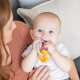 A baby with big blue eyes chews on Bumkins Silicone Teething Charms: Orange, offering teething relief, held by a brown-haired woman.