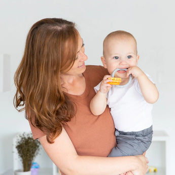 A woman holds a baby with Bumkins Silicone Teething Charms: Orange, offering teething relief in a bright room.