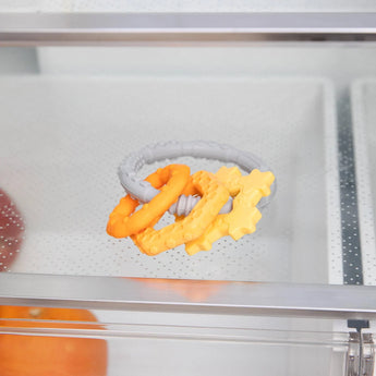 Bumkins Silicone Teething Charms in orange with star textures, made of food-safe silicone, offer teething relief on a glass shelf.