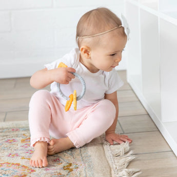 A baby in a white bow headband sits on a rug with Bumkins Orange Silicone Teething Charms, offering teething relief by white shelves.