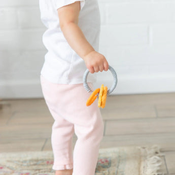 A toddler in a white shirt and pink pants holds Bumkins Silicone Teething Charms: Orange, standing on a rug in a bright room.