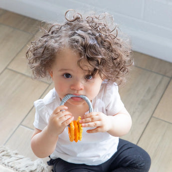 Toddler with curly hair in a white shirt and black pants holds a Bumkins Silicone Teething Charms: Orange near their mouth.