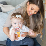 On a cozy couch, a woman smiles as her baby enjoys chewing on Bumkins Silicone Teething Charms: Orange while sporting a bow headband.