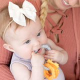 A baby with a white bow chews on a Bumkins Silicone Teething Charm: Orange, held by someone in a peach sweater.