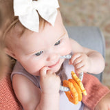 Baby in a purple outfit, wearing a white bow, enjoys teething relief with Bumkins Orange Silicone Teething Charm on someones lap.