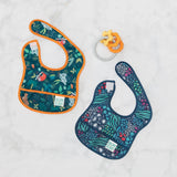 Two colorful baby bibs rest on a white surface: one with a jungle animal pattern and orange border, the other with a dark floral leaf design. Between them lies Bumkins Silicone Teething Charms in orange, featuring vibrant beads for teething relief.