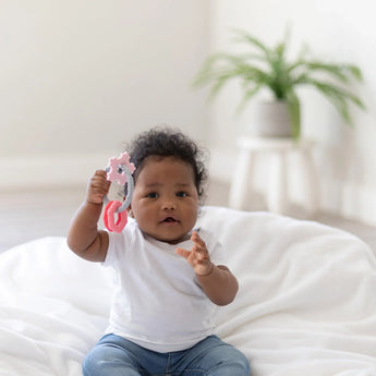 Baby in a white tee and jeans sits on a soft blanket holding Bumkins Silicone Teething Charm: Pink, with a potted plant behind.