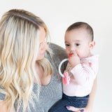 A woman with long blonde hair holds a baby in pink chewing on Bumkins Silicone Teething Charms: Pink to aid motor skill development.