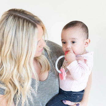 A woman with long blonde hair holds a baby in pink chewing on Bumkins Silicone Teething Charms: Pink to aid motor skill development.