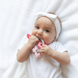 Baby in white outfit with headband chews on Bumkins Silicone Teething Charms: Pink, enhancing motor skills.