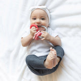 Baby on a white blanket, smiling softly, holding Bumkins Silicone Teething Charms: Pink and honing motor skills with a headband on.