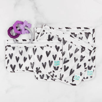 Four Bumkins heart-patterned bags on a marble surface with purple 100% food-safe Silicone Teething Charms.