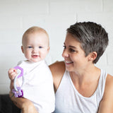 A short-haired woman smiles at a baby with a Bumkins Silicone Teething Charm: Purple; both wear white tops in a bright room.