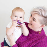 A smiling, short-haired person in a purple sweater and earrings holds a laughing baby in a white shirt chewing on Bumkins Silicone Teething Charms: Purple, gazing affectionately at the baby against a plain background.