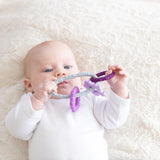 A baby in a white onesie chews on Bumkins 100% food-safe Silicone Teething Charms: Purple for relief on a fluffy blanket.