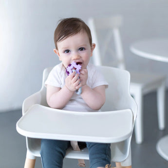 Baby in a high chair chewing on Bumkins Silicone Teething Charms: Purple, with a white table and chair in the background.