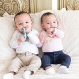 Two babies on a couch chew on Bumkins food-safe Silicone Teething Charms; one wears white, the other sports a pink bib over a white top.