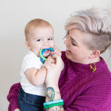 A smiling adult with short blonde hair holds a baby chewing on Bumkins Silicone Teething Rings from the Summer 4 Pack, wearing a purple sweater.