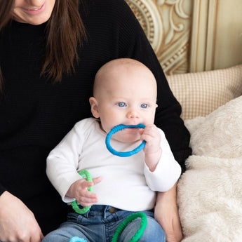 A baby in a white shirt holds a blue Bumkins Silicone Teething Ring, sitting on an adults lap in a cozy room.