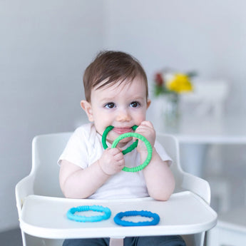 A baby in a high chair chews on Bumkins Silicone Teething Rings from the Summer pack, with blue rings visible. Flowers bloom blurred in back.