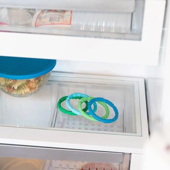 Bumkins Silicone Teething Rings 4 Pack: Summer sit on a glass fridge shelf, with a covered dish of food above.