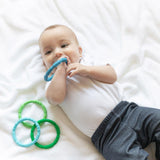 Baby lying on a white blanket, wearing a white onesie and gray pants, chewing Bumkins silicone teething ring from the Summer pack.