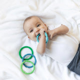 Baby on a white blanket chewing a Bumkins Summer Silicone Teething Ring, with green and blue rings nearby.