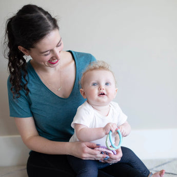 A woman smiles at a blue-eyed baby with a Bumkins Silicone Teething Ring from the Spring pack, both on the floor against a light wall.