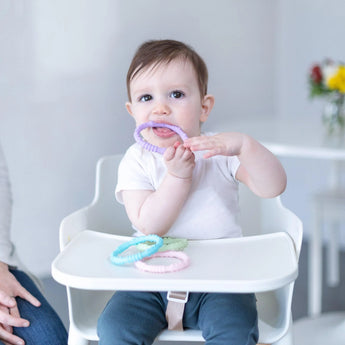 A baby in a high chair chews on a Bumkins pastel purple silicone teething ring from the Spring 4 Pack, surrounded by other toys.