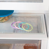 The Silicone Teething Rings 4 Pack from Bumkins are pastel-colored and sit beside a blue-lidded bowl on a glass fridge shelf.