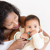 A smiling woman holds a baby, dressed lightly, chewing on Bumkins Silicone Teething Ring to soothe sore gums.