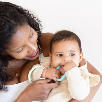 A smiling woman holds a baby, dressed lightly, chewing on Bumkins Silicone Teething Ring to soothe sore gums.