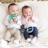 Two babies sit on a plush, cream-colored blanket with colorful Bumkins Silicone Teething Rings. The baby on the left wears a white shirt and light pants, while the other is dressed in a pink top and jeans.