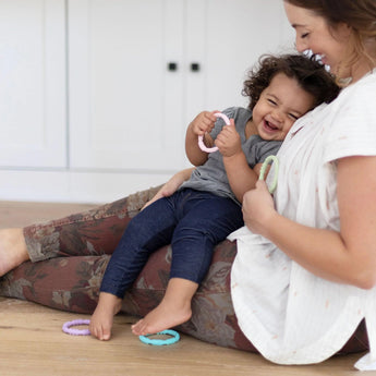 A woman sits with a laughing toddler in a cozy home, both enjoying Bumkins Silicone Teething Rings 4 Pack: Spring.