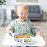 A toddler in a high chair enjoys spaghetti, blueberries, and broccoli on a divided plate, wearing a Bumkins Sleeved Bib: Cacti.