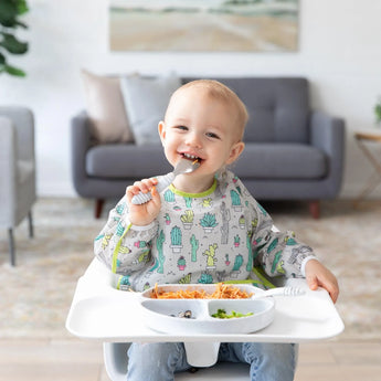 A happy toddler in a highchair enjoys spaghetti and peas using a spoon while wearing Bumkins Sleeved Bib: Cacti.