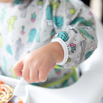 A child wears a Bumkins Sleeved Bib: Cacti, made from waterproof fabric, at the table with a spoon ready above a bowl.