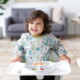 A toddler in a Bumkins Sleeved Bib: Cacti enjoys spaghetti in a high chair. A sofa and side table are visible in the background.