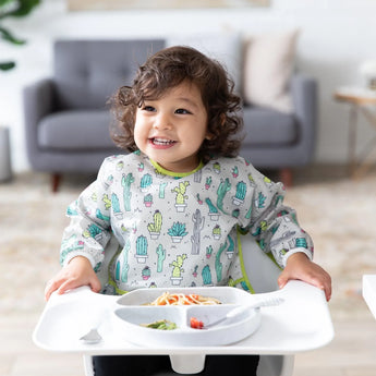 A toddler in a Bumkins Sleeved Bib: Cacti enjoys spaghetti in a high chair. A sofa and side table are visible in the background.