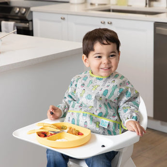 A smiling toddler in a high chair wears a Bumkins Sleeved Bib: Cacti and enjoys food on a divided yellow plate. Kitchen background.