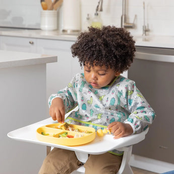 A curly-haired child enjoys food from a yellow divided plate, wearing a Bumkins Sleeved Bib: Cacti for a perfect fit in the kitchen.