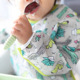 A baby holds a food-covered spoon, wearing Bumkins Sleeved Bib: Cacti with adjustable fit and fun waterproof fabric.