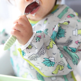 A baby holds a food-covered spoon, wearing Bumkins Sleeved Bib: Cacti with adjustable fit and fun waterproof fabric.