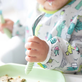 A baby enjoys a meal mess-free with Bumkins Sleeved Bib: Cacti, using a spoon and sitting at the table with a green plate.