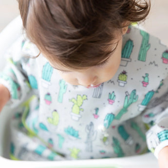 A toddler in a Bumkins Sleeved Bib: Cacti sits in a high chair, glancing down with soft, curly hair.