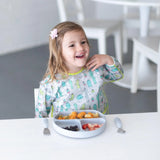 A smiling child wearing a Bumkins Sleeved Bib: Cacti sits at the table with fruit, pasta, and a flower hair clip.