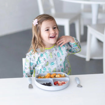 A smiling child wearing a Bumkins Sleeved Bib: Cacti sits at the table with fruit, pasta, and a flower hair clip.