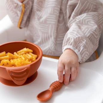 A child in a Bumkins Sleeved Bib: Wander with Desert Boho patterns reaches for a spoon beside their bowl of mac and cheese.