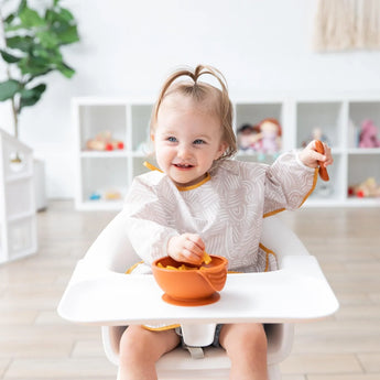 A toddler wearing a Bumkins Sleeved Bib: Wander smiles while eating from an orange bowl; Desert Boho toys and decor fill the shelves.