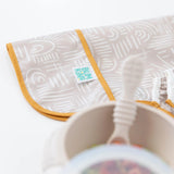 Close-up of Sleeved Bib: Wander in beige Desert Boho fabric with Bumkins label. Blurred bowl and spoon in the foreground.